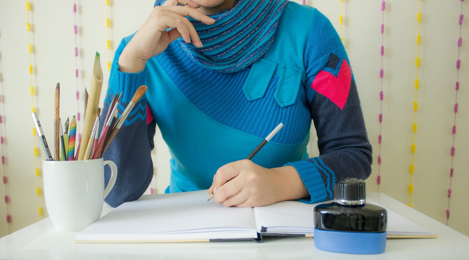 Person writing in a notebook beside a cup of pencils and brushes